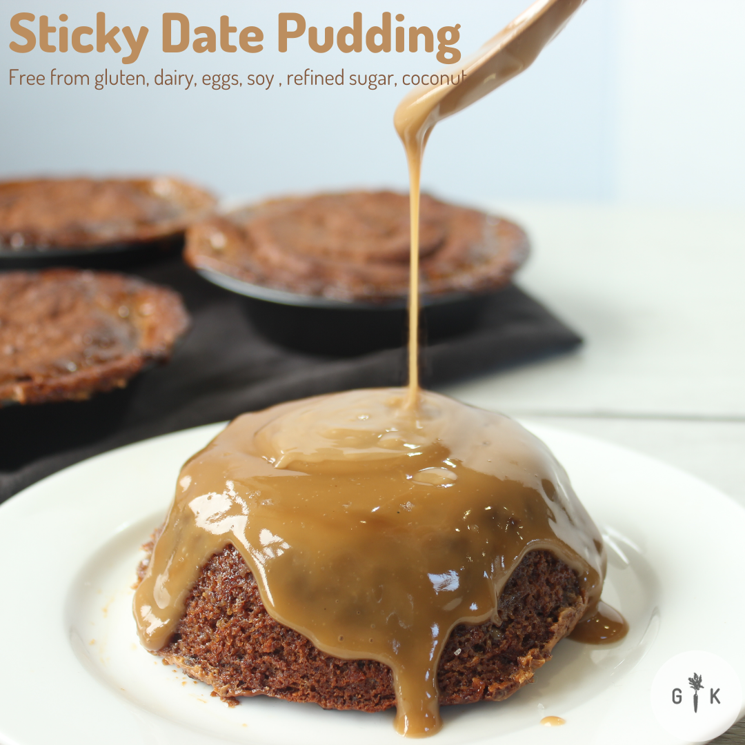 Gluten free sticky date pudding on a white plate in the foreground, a gold spoon drizzling caramel sauce over the sticky date pudding. 3 sticky date pudding serves in the background.